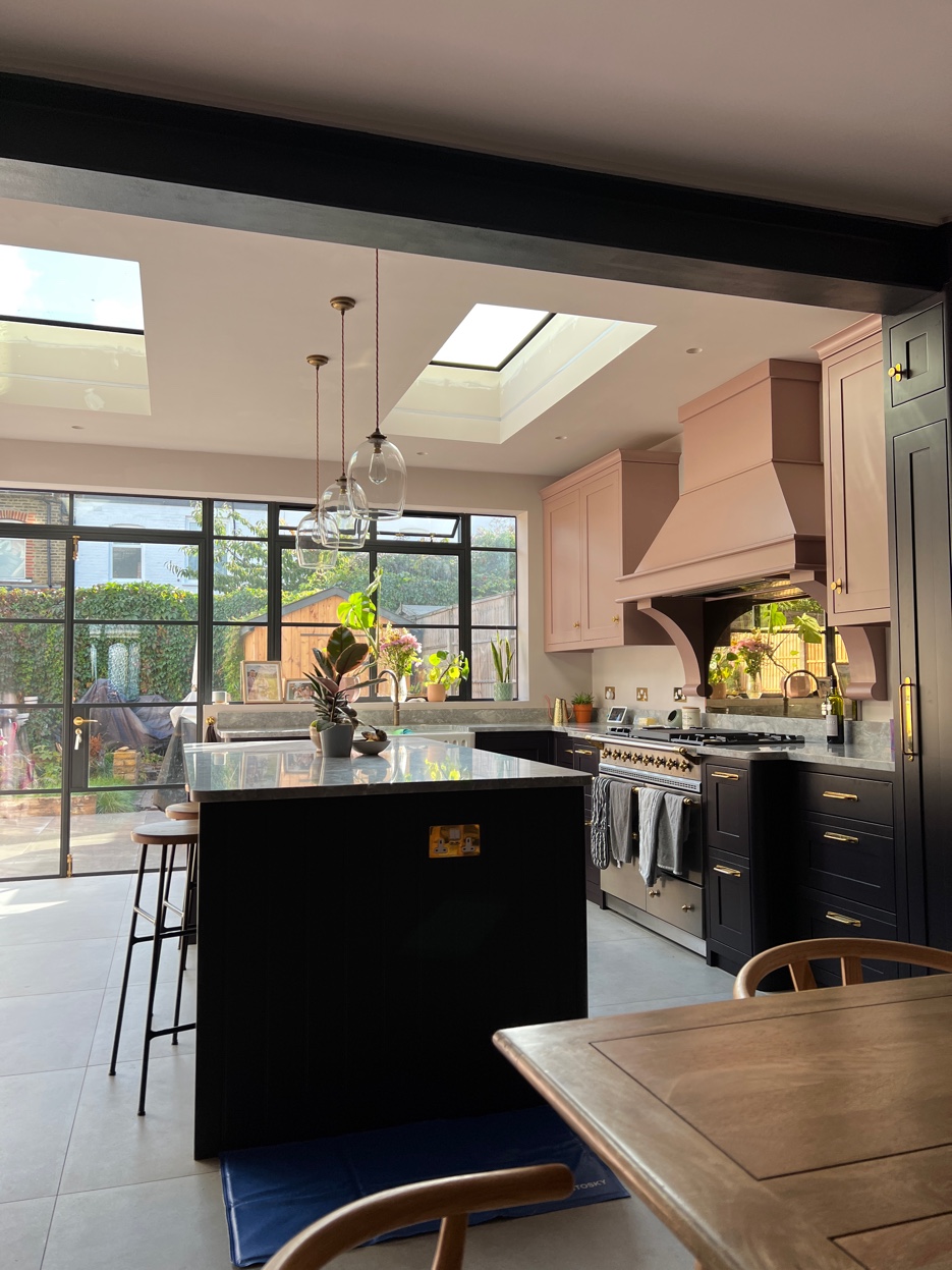 Wide kitchen view from the dining area showing dark lower cabinets, dusty pink uppers, quartzite island, and crittall-style glazing onto the garden