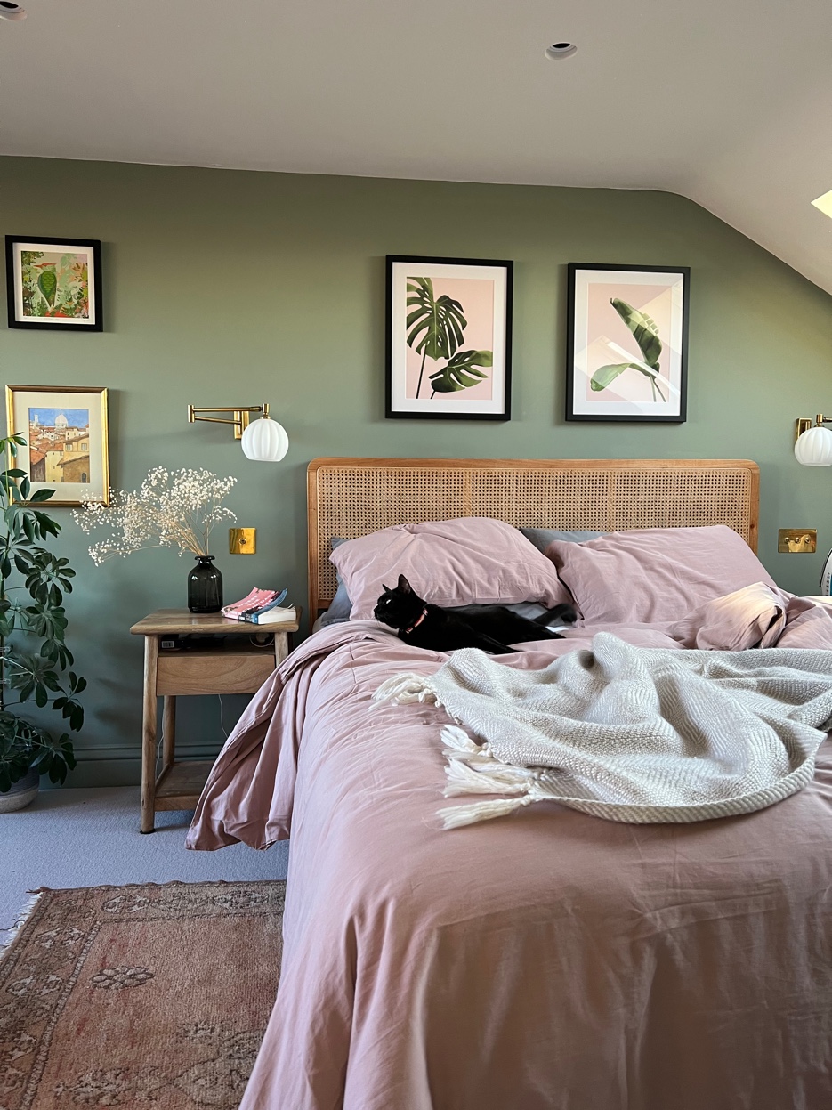 Loft bedroom in sage green with rattan headboard, dusty pink linen, botanical prints, and a cat on the bed